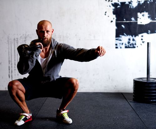 Athlete in a squat hold with dumbbells in a gym, wearing dark workout clothes and colorful trainers.