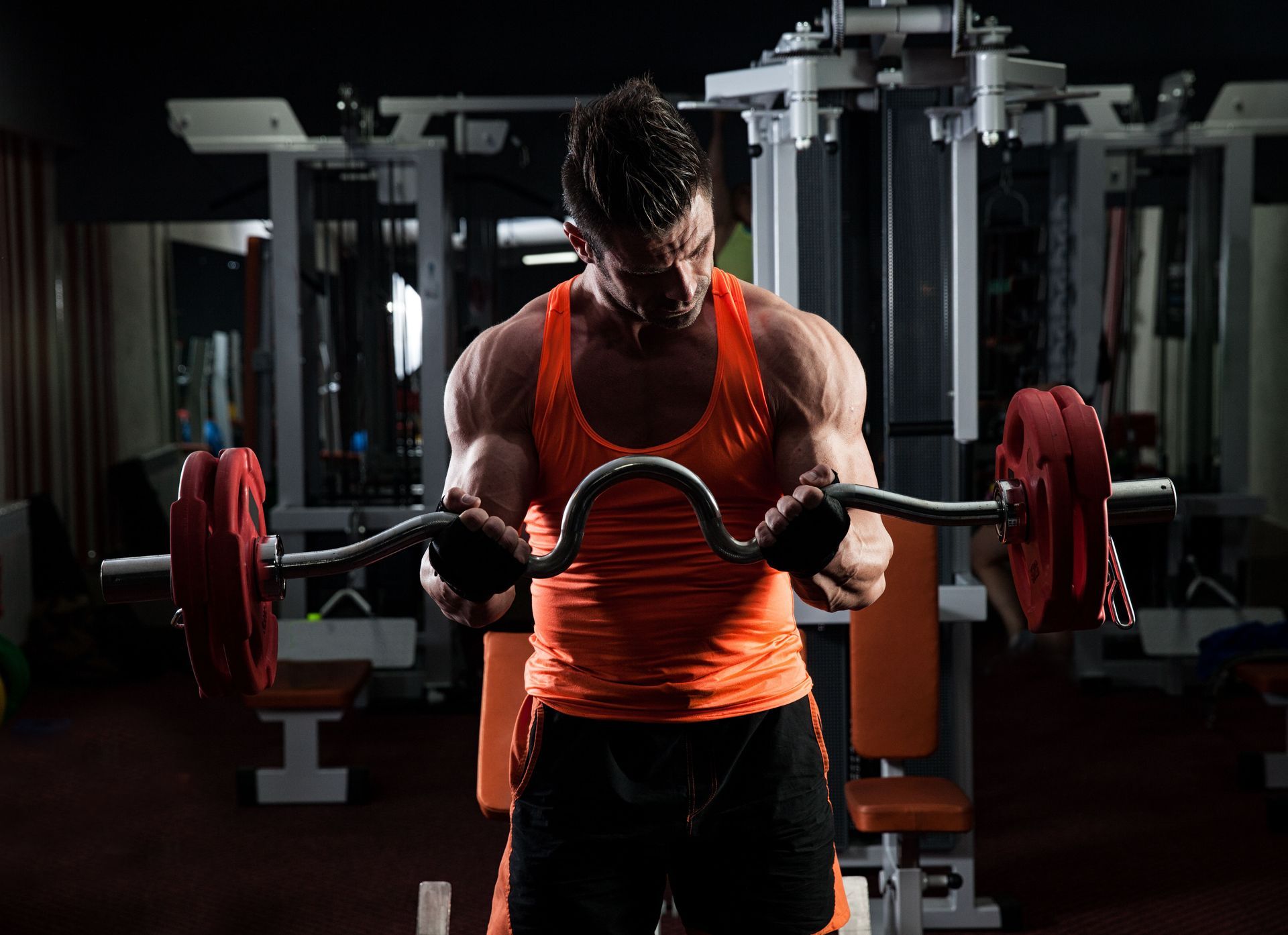 Man lifting a barbell in a dark gym, wearing an orange tank top, focused on a curl exercise.