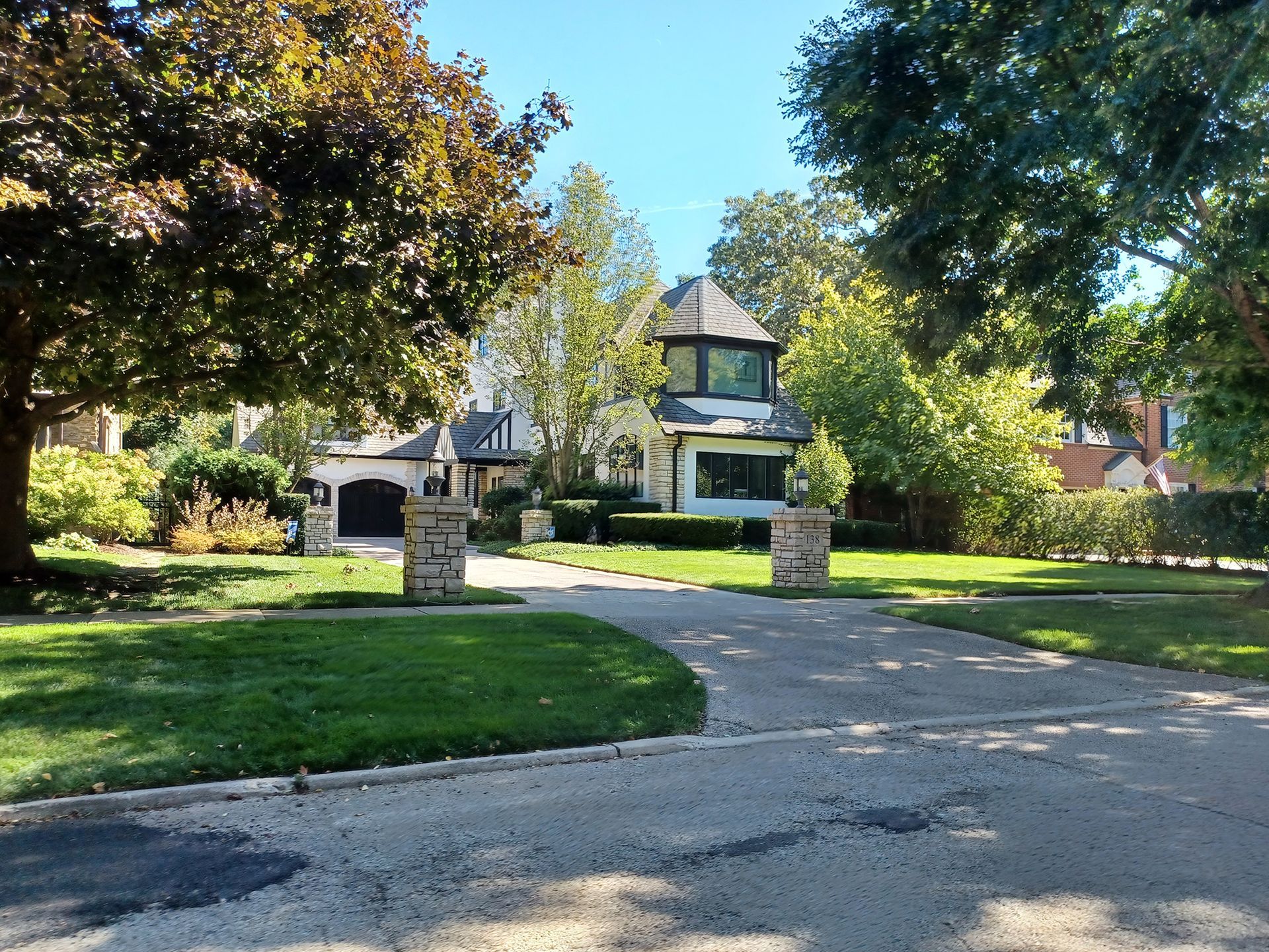 House with turret at the end of a curved driveway, flanked by stone pillars and trees on a sunny day.