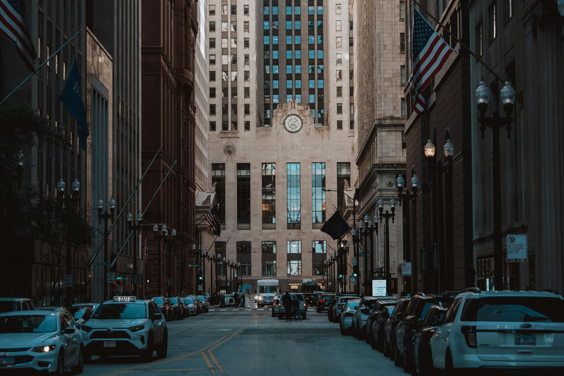 Street view of tall buildings, cars parked along the sides, leading to a prominent building with a clock.