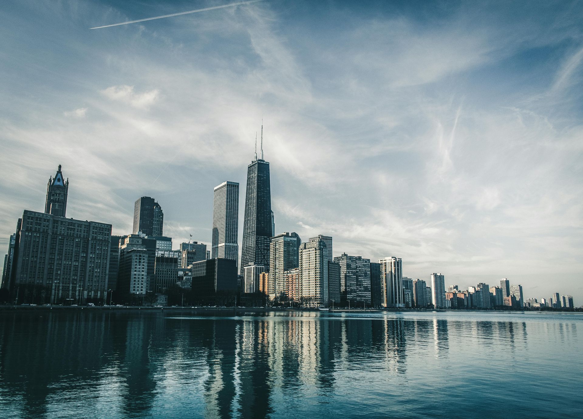Chicago skyline reflecting in calm water under a blue and cloudy sky.