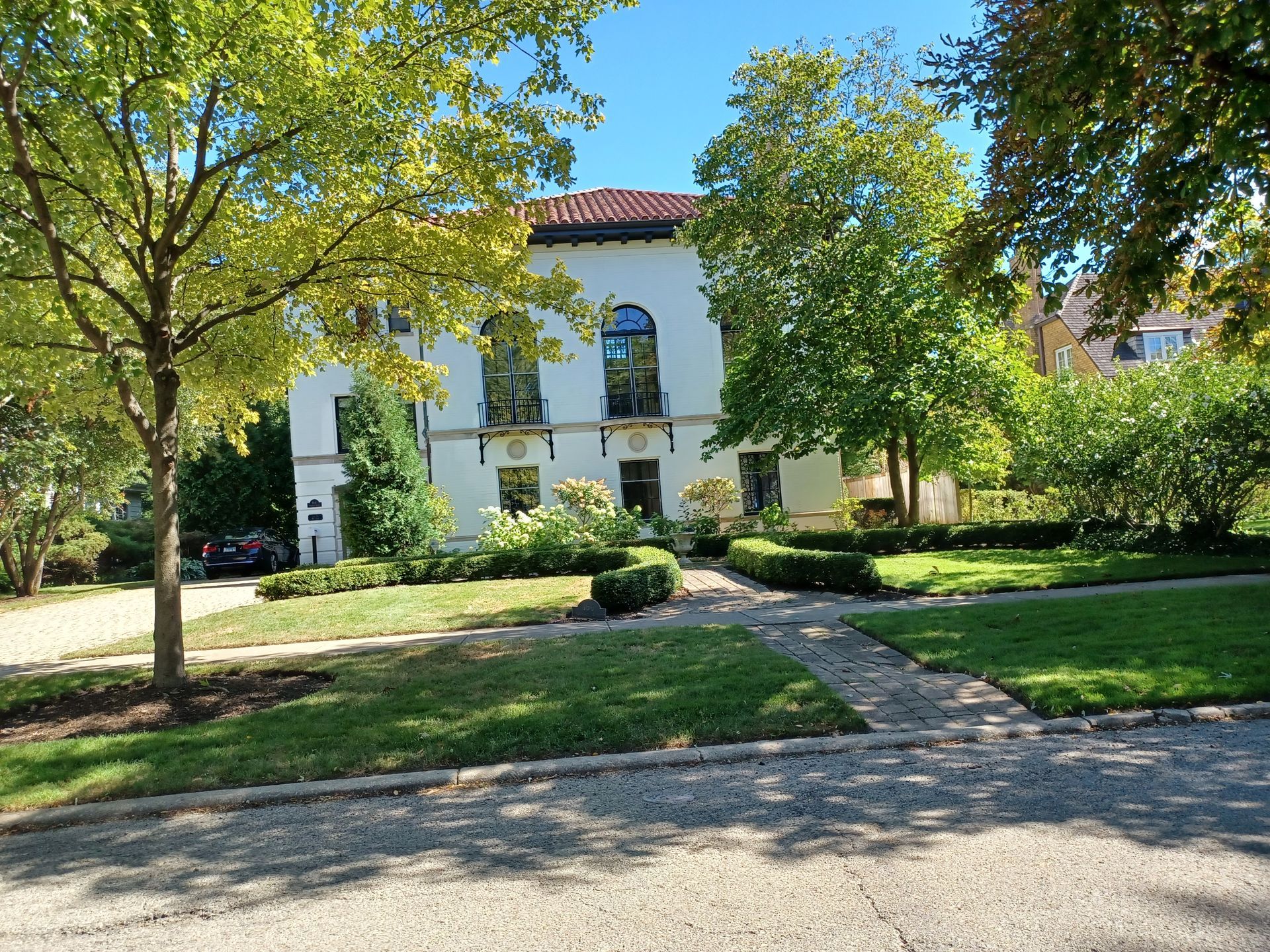 White stucco building with arched windows, red tile roof, and manicured lawn. Trees frame the view.