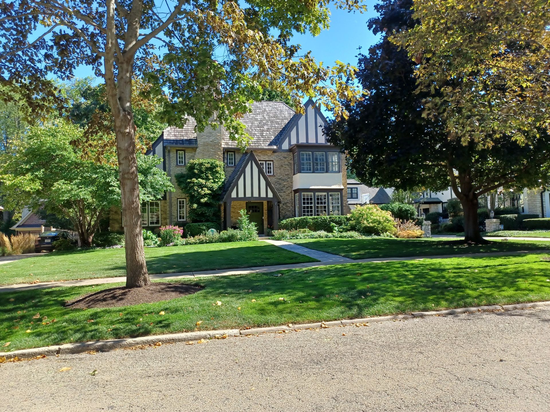 Tudor-style house with stone and timber facade, surrounded by trees and a green lawn on a sunny day.