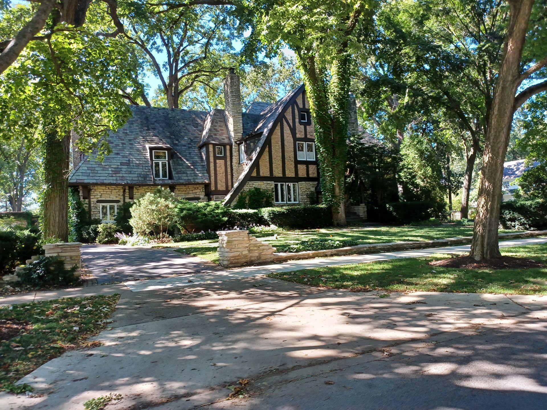 Tudor-style house with stone and brick exterior, set among large trees. Driveway leads to the house on a sunny day.