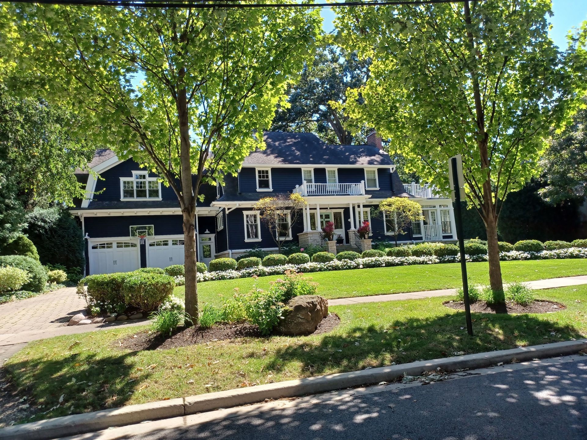 Dark blue house with white trim, two-car garage, and a porch. Trees and bushes in the yard.