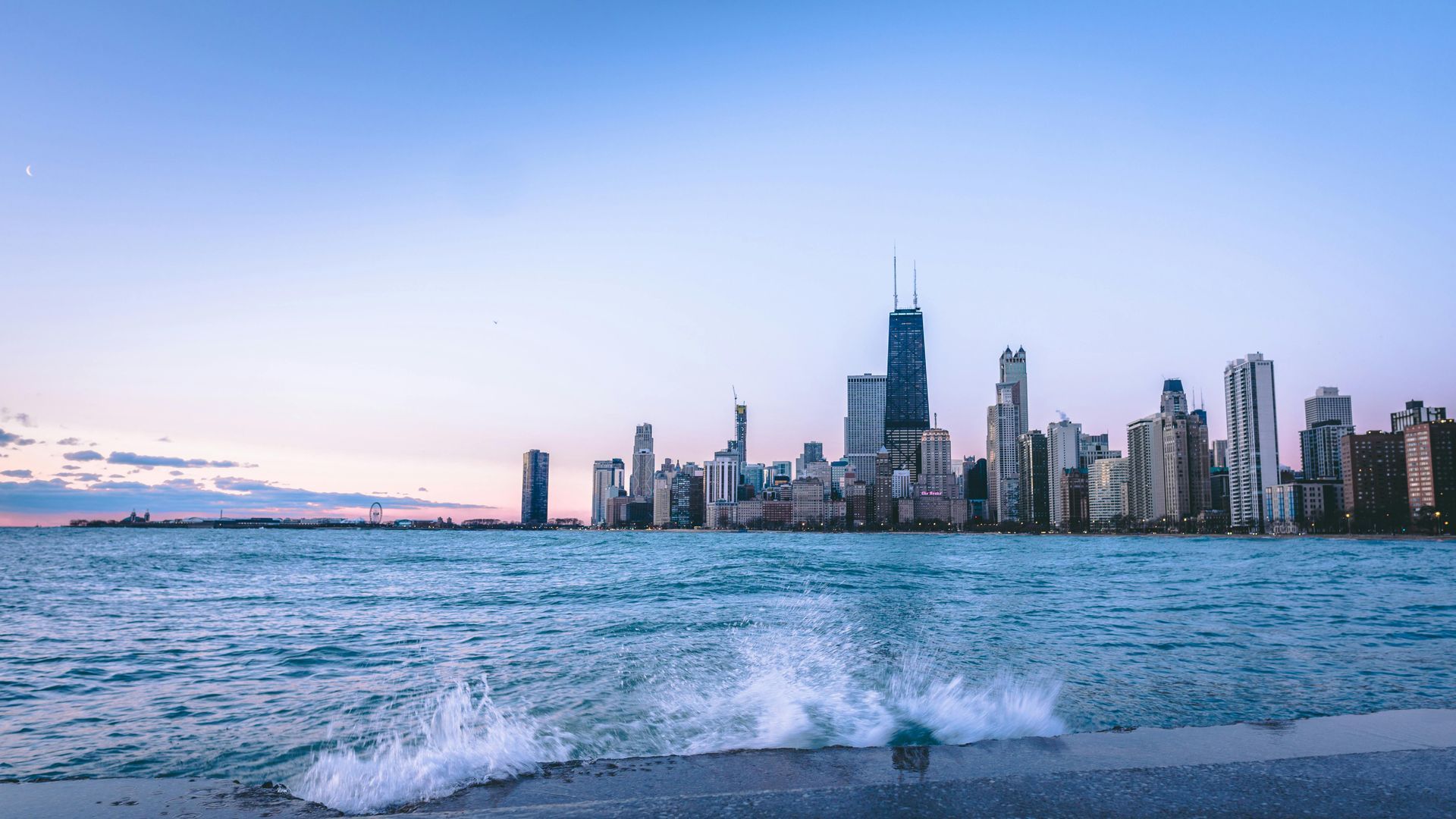 City skyline across choppy water at dusk, with waves splashing in the foreground and a pastel sky.