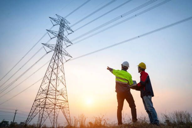 Two workers in safety vests examine a power line tower at sunset.