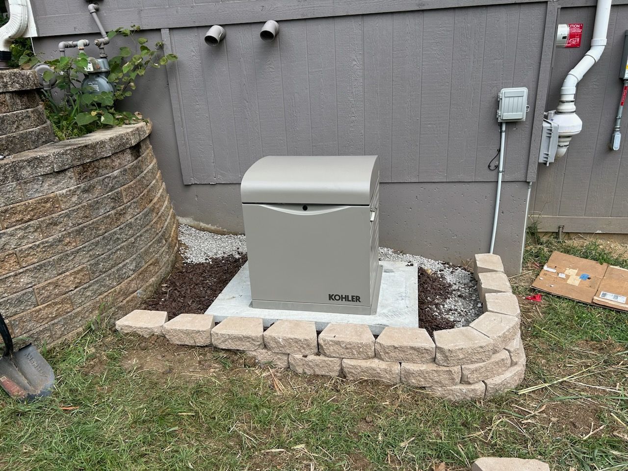 A gray generator on a concrete pad surrounded by a brick border and mulch, set against a gray house.