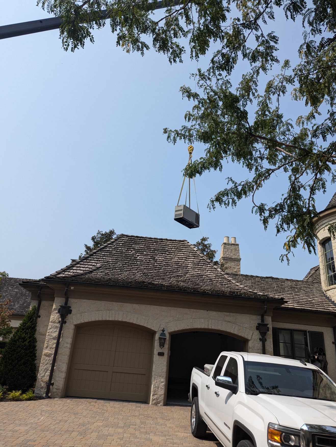 A crane lifting an air conditioning unit above a house with a tiled roof. A white truck is parked nearby.