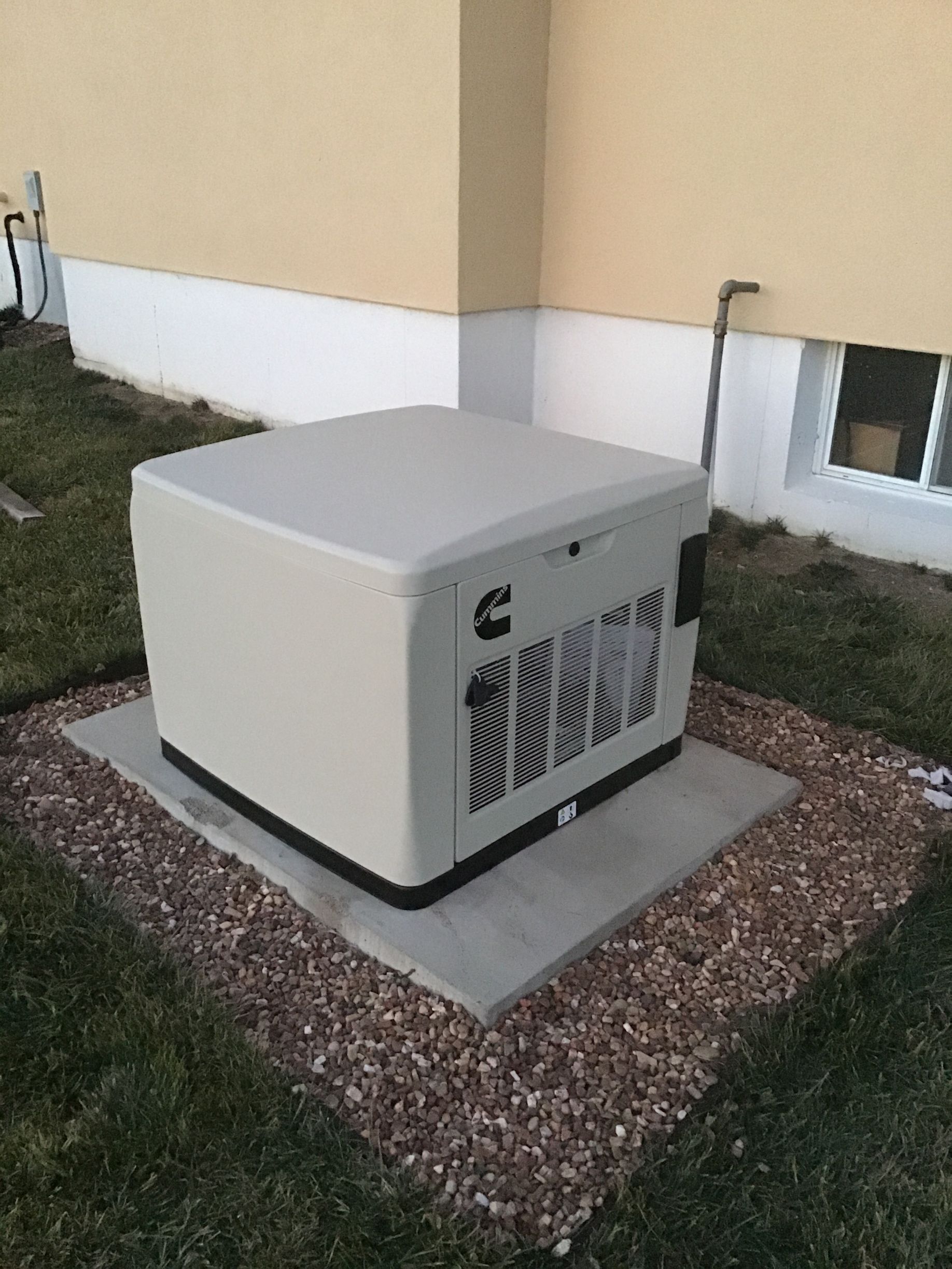 Generator outside a building, cream-colored with black accents, sits on a concrete pad surrounded by gravel and grass.