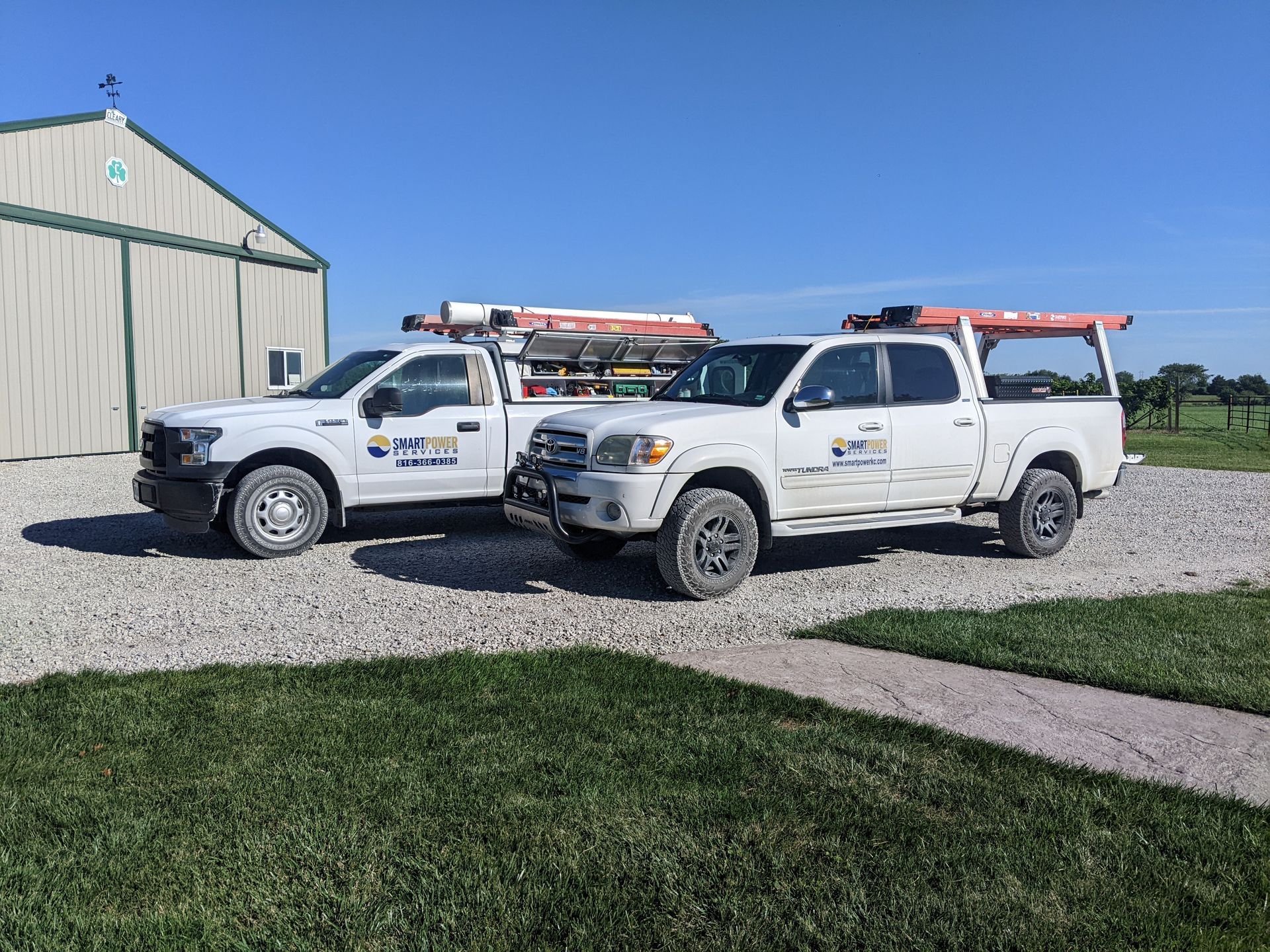 Two white pickup trucks parked on gravel in front of a building on a sunny day.