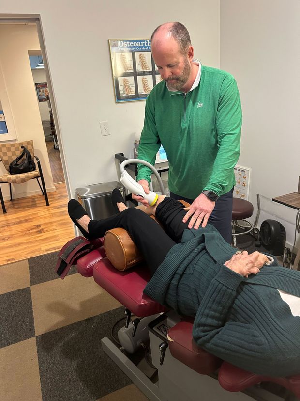Man in green shirt using a device on a patient's leg while she lies on an examination table.