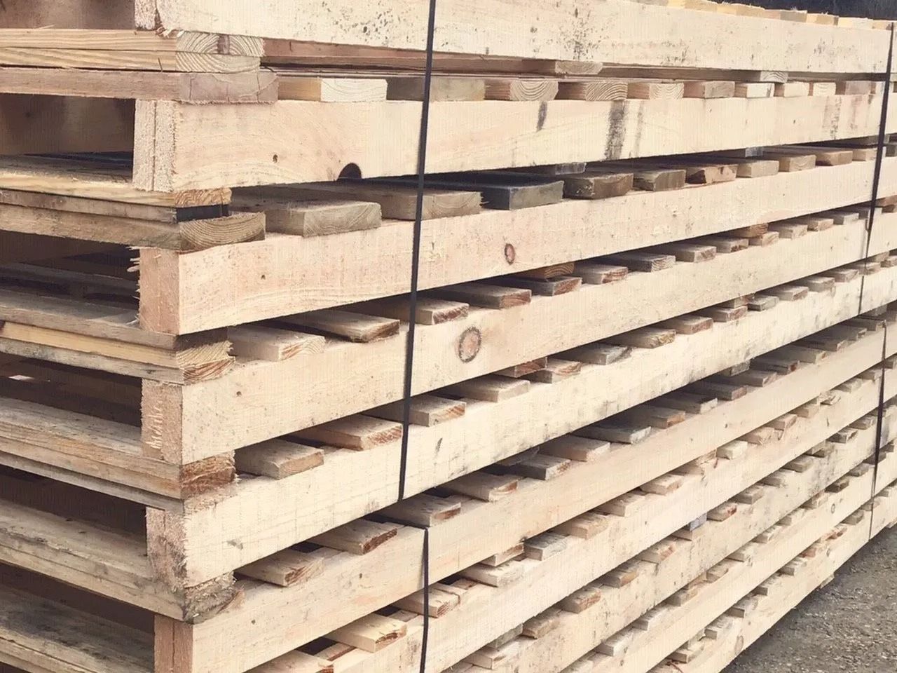 A stack of light-colored wooden shipping pallets, secured with black straps, viewed from a slightly angled perspective.