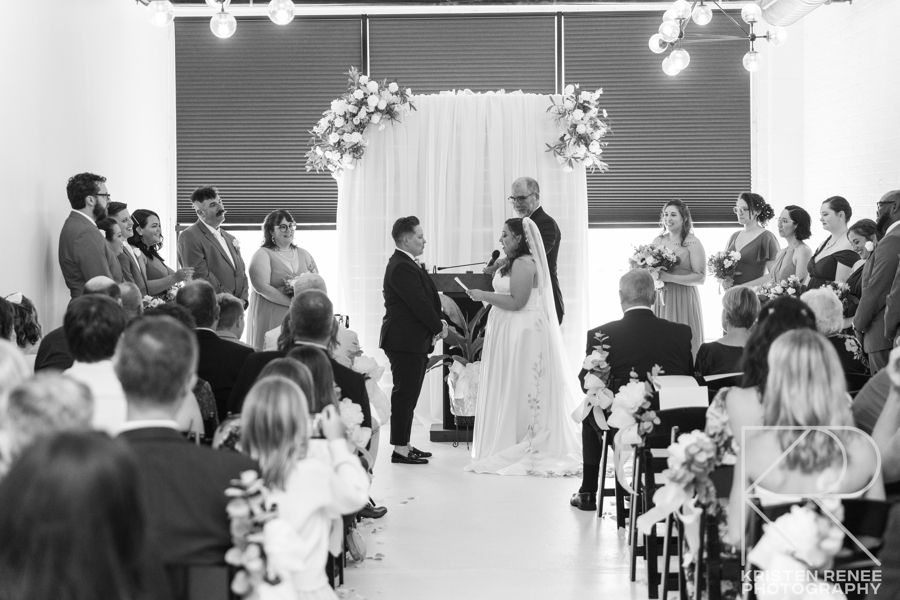 A black and white photo of a bride and groom holding hands during their wedding ceremony.