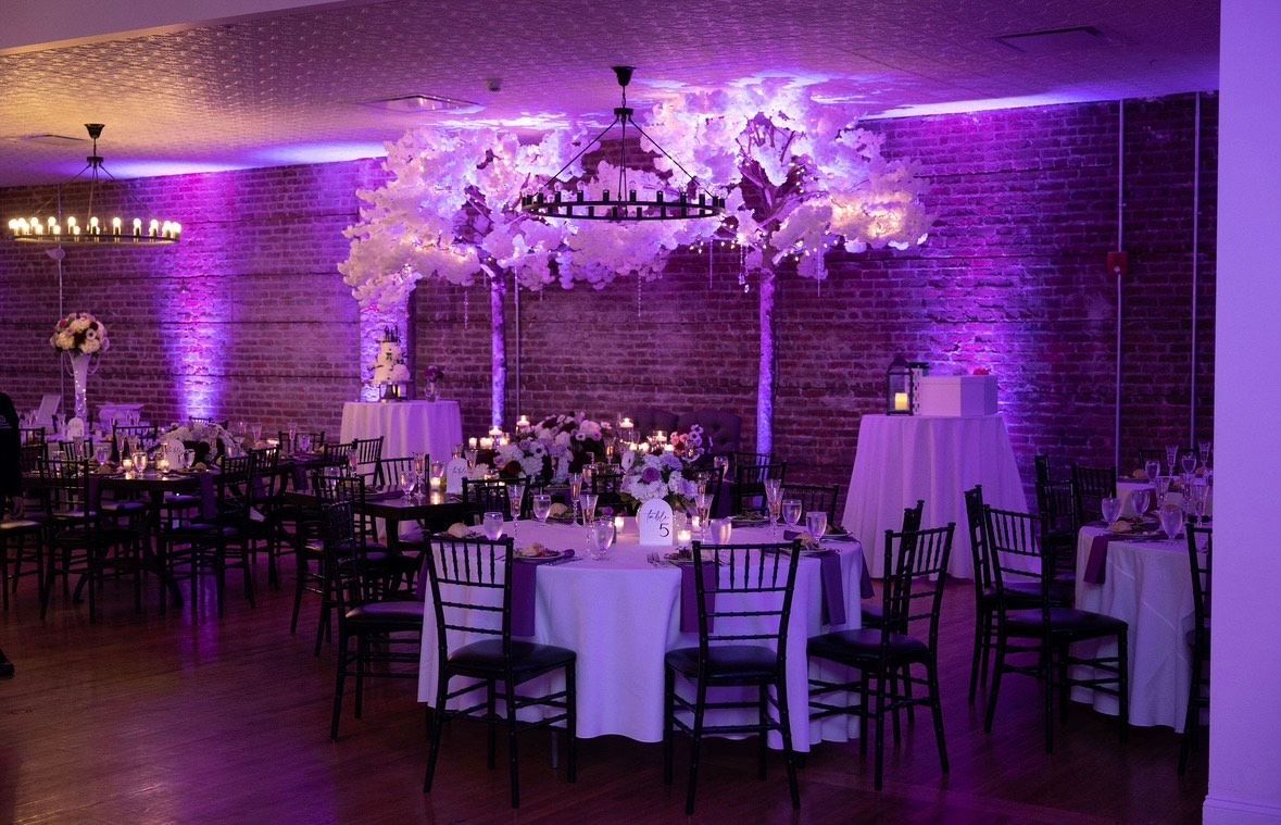 A large room with tables and chairs set up for a wedding reception with purple lighting.