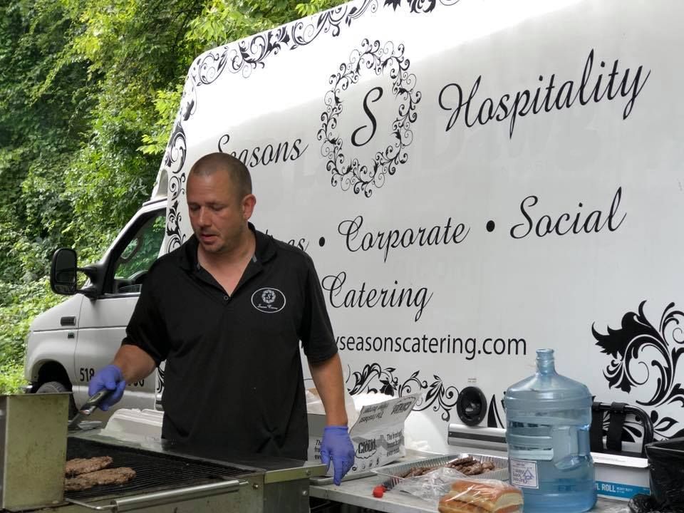 A man is grilling food in front of a white van that says seasons hospitality