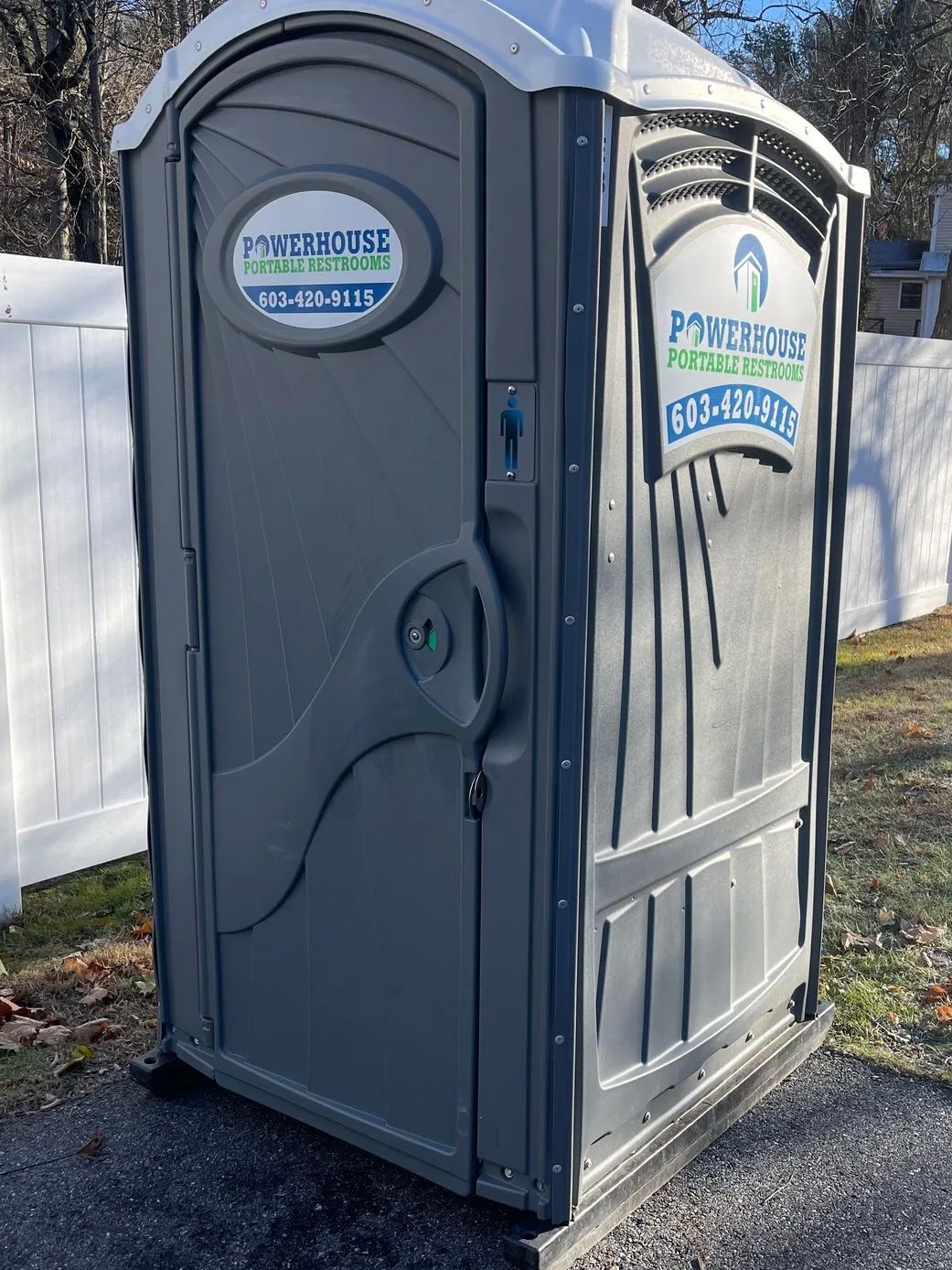 A grey portable restroom unit from P-Title House standing outdoors against a white fence.