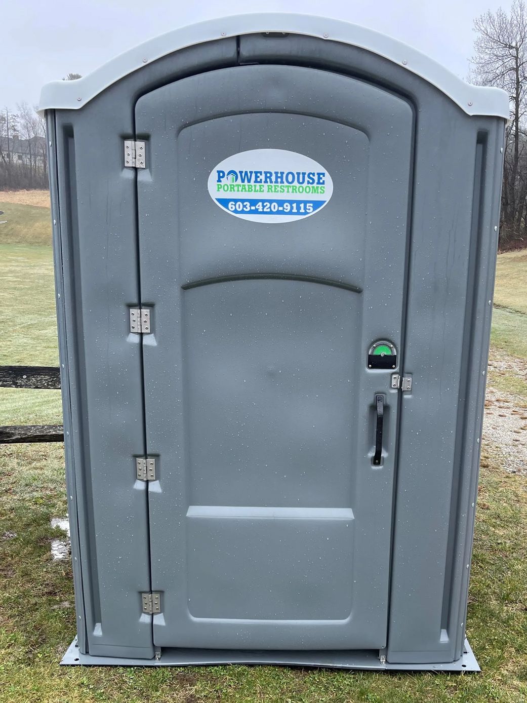 A grey portable toilet stands on a grassy field, featuring a white logo with contact information on the door.
