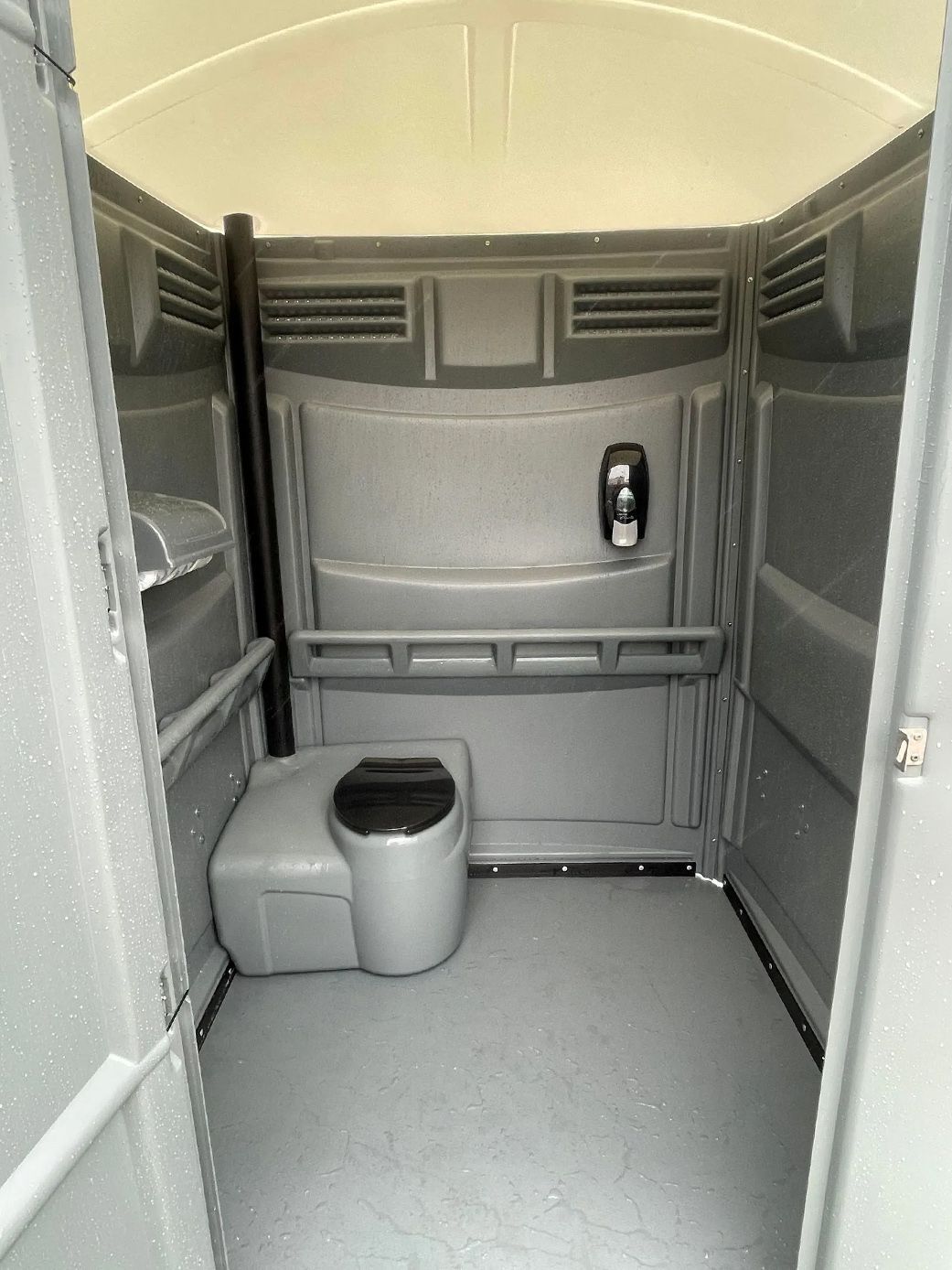 Interior view of a gray portable toilet featuring a floor-mounted toilet seat, a wall-mounted hand sanitizer dispenser.