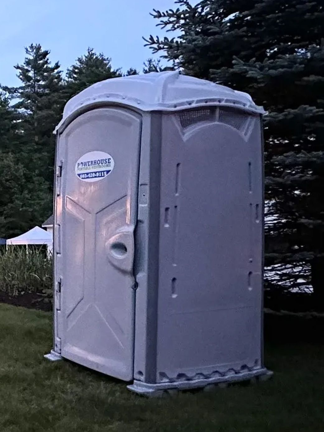 A light gray portable toilet standing on a grass lawn near some pine trees.
