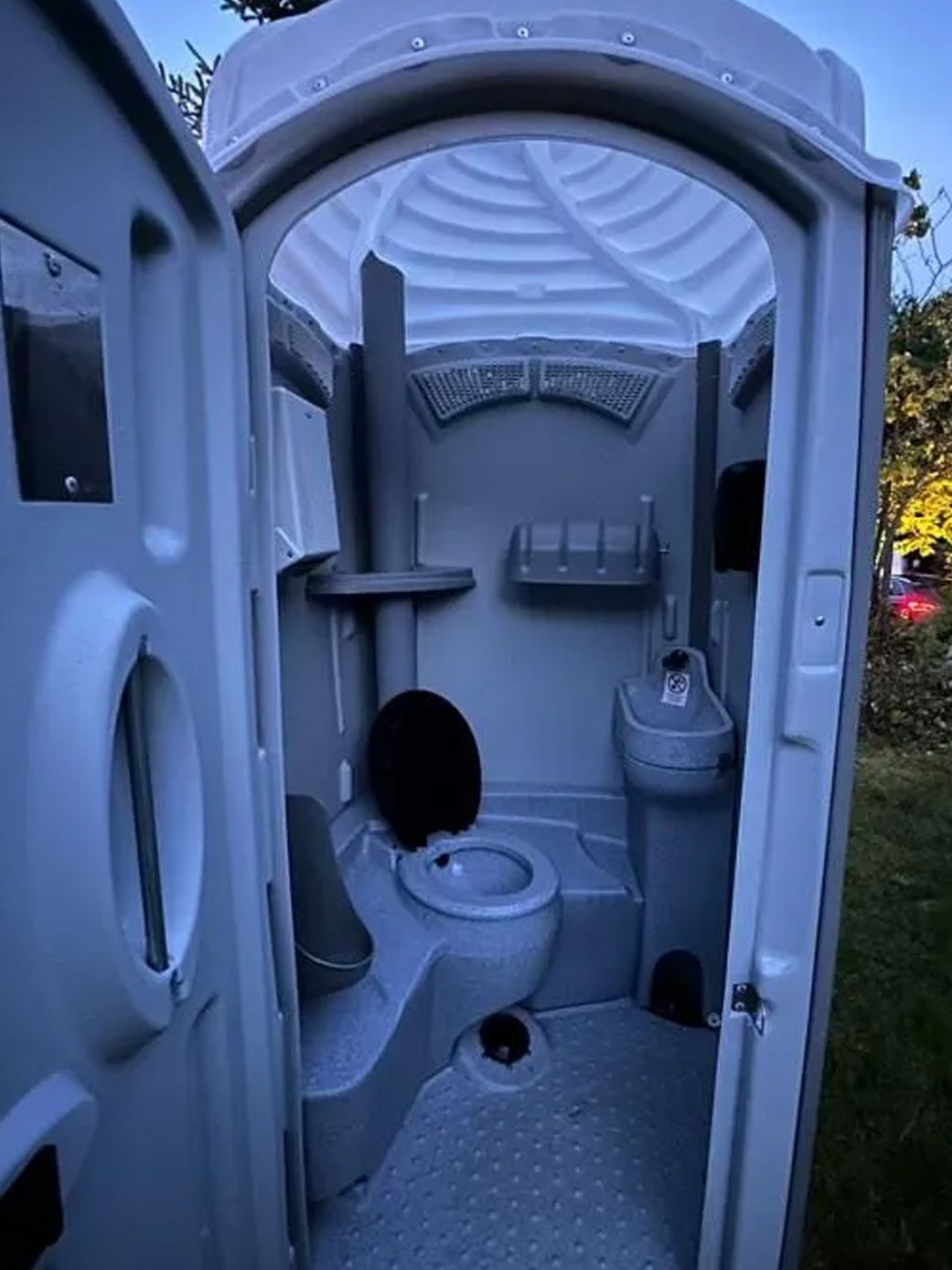 An interior view of a grey portable toilet with a sink, black toilet seat, and shelf, standing outdoors at twilight.