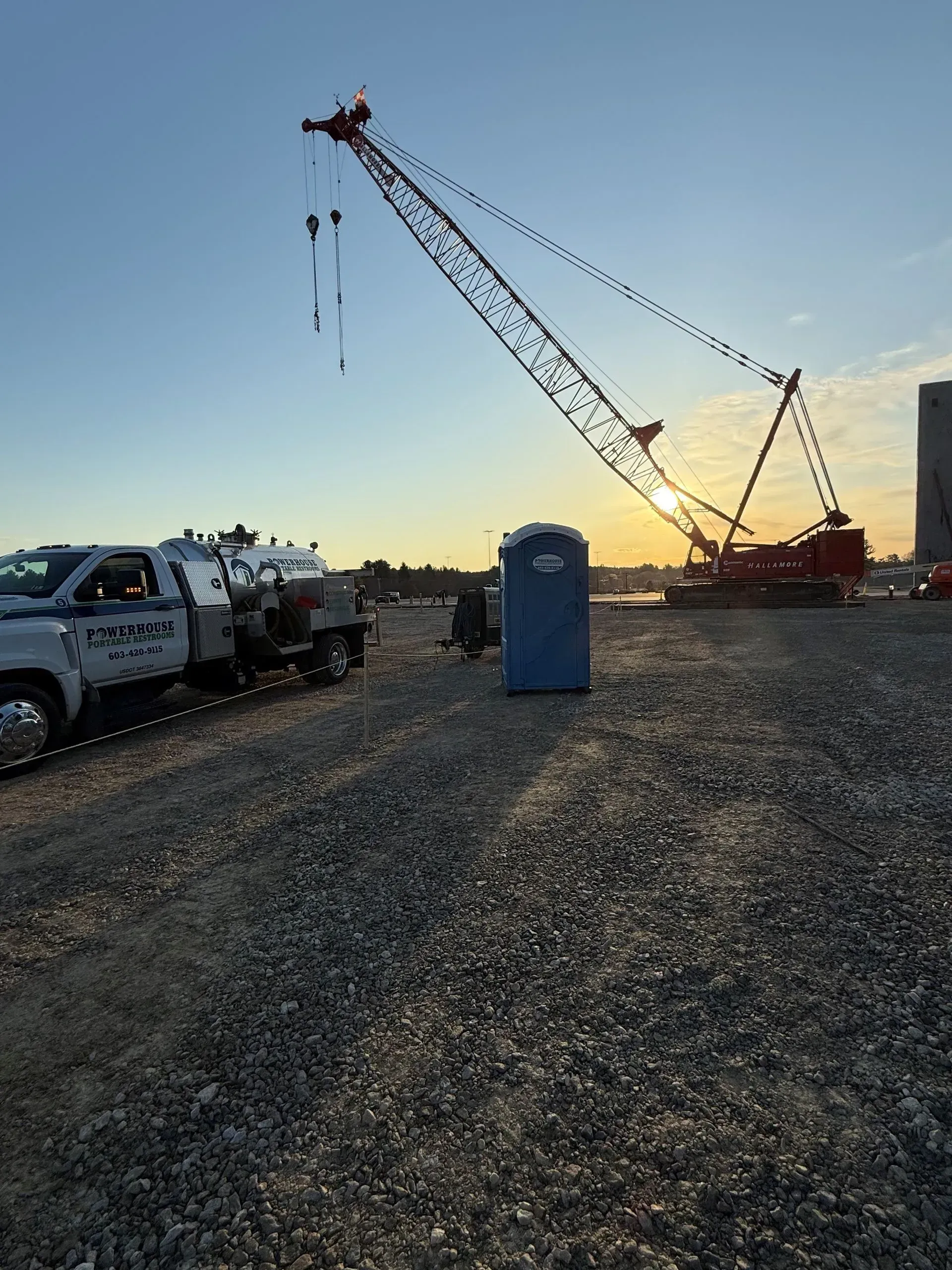 A large crane towers over a gravel construction site at sunrise, with a service truck and portable toilet nearby.