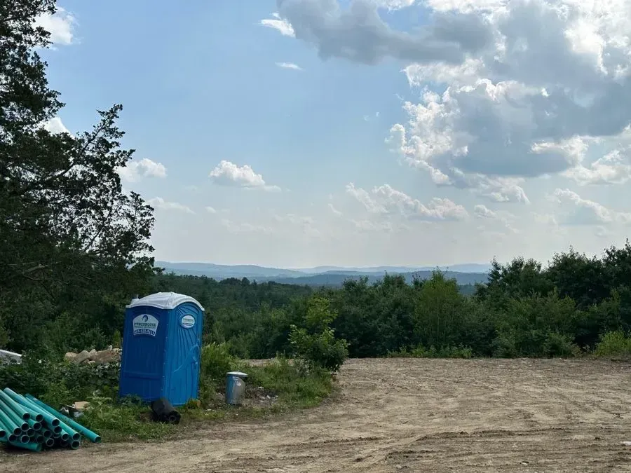 A portable toilet and a stack of green pipes sit at the edge of a cleared dirt lot overlooking a hazy, forested valley.