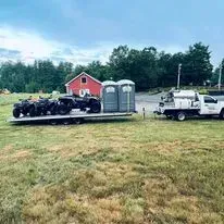 A trailer carrying four ATVs and two portable toilets is parked in a grassy field in front of a red building and a truck.