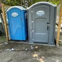 Two portable toilets, one blue and one gray, sit side-by-side on a wooden platform against a lattice fence outdoors.