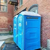 Two blue portable toilets standing on an asphalt surface against a brick building wall.