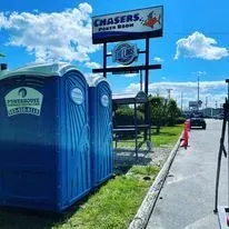 Two blue portable toilets stand in front of a Chasers Sports Bar sign under a sunny, partly cloudy sky.