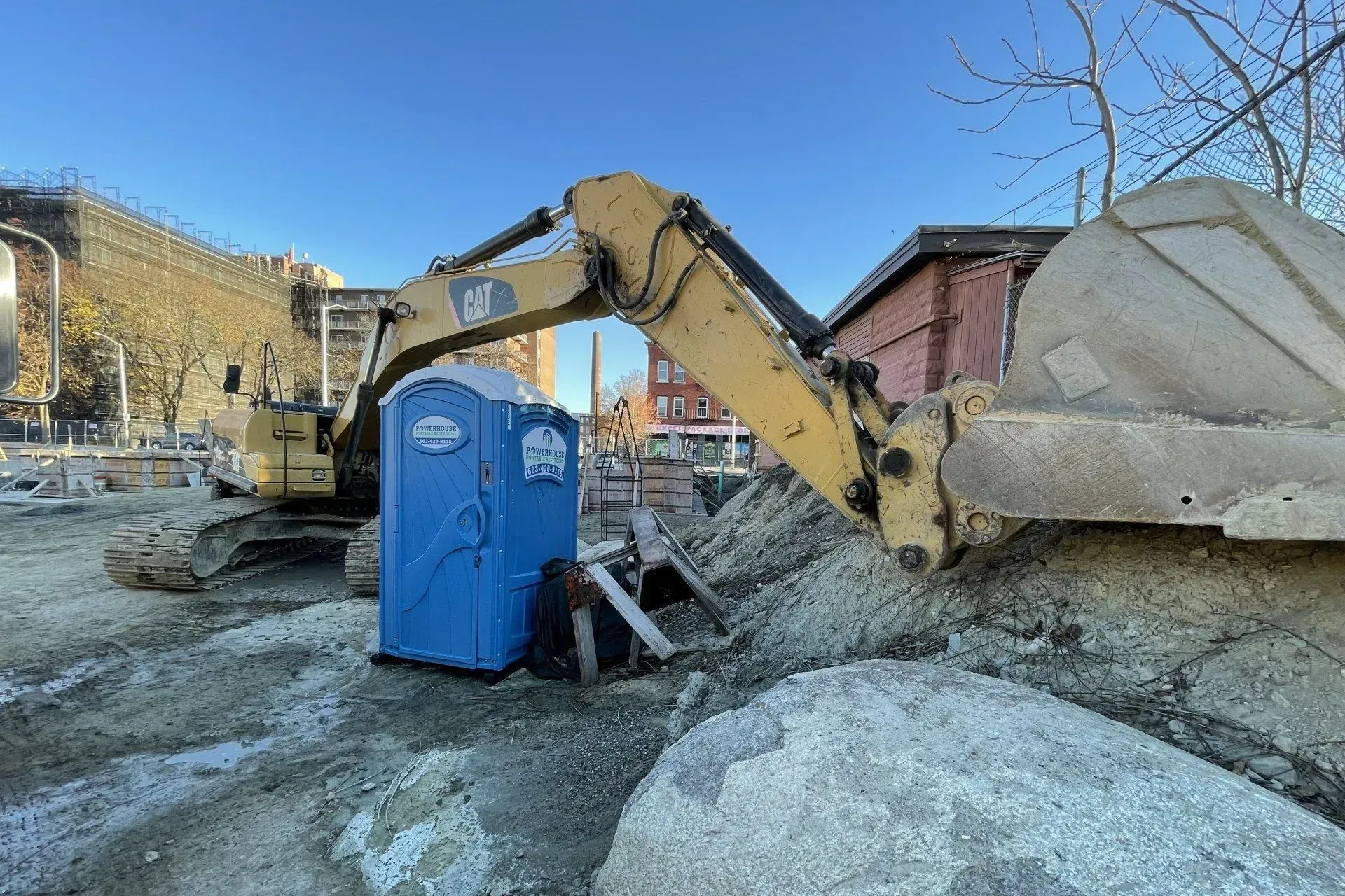 A yellow excavator parked on a dirt construction site next to a bright blue portable toilet under a clear blue sky.