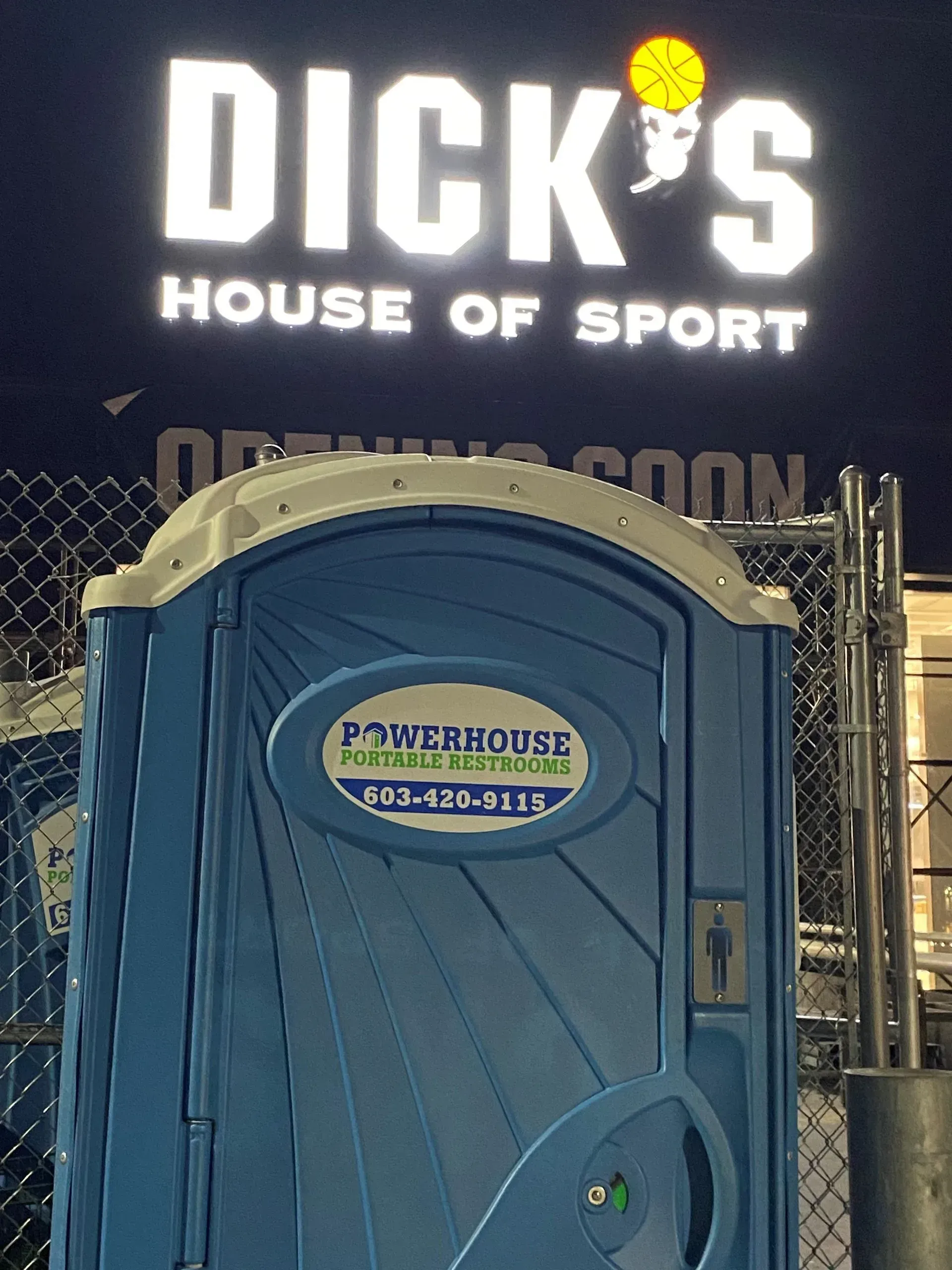 A blue portable restroom sits in front of a brightly lit 