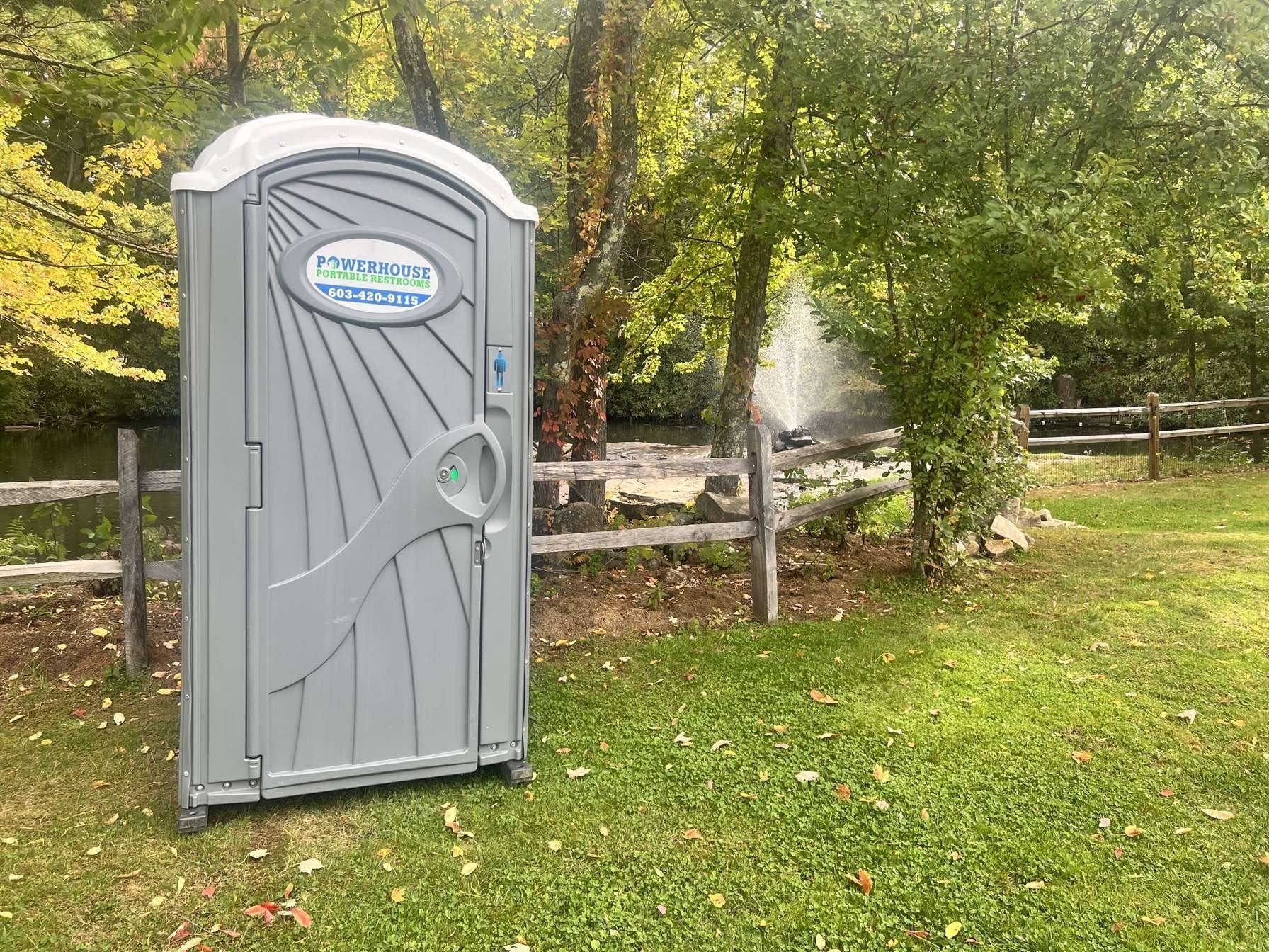 A gray portable toilet sits on a grassy lawn next to a wooden fence and a fountain in a park.