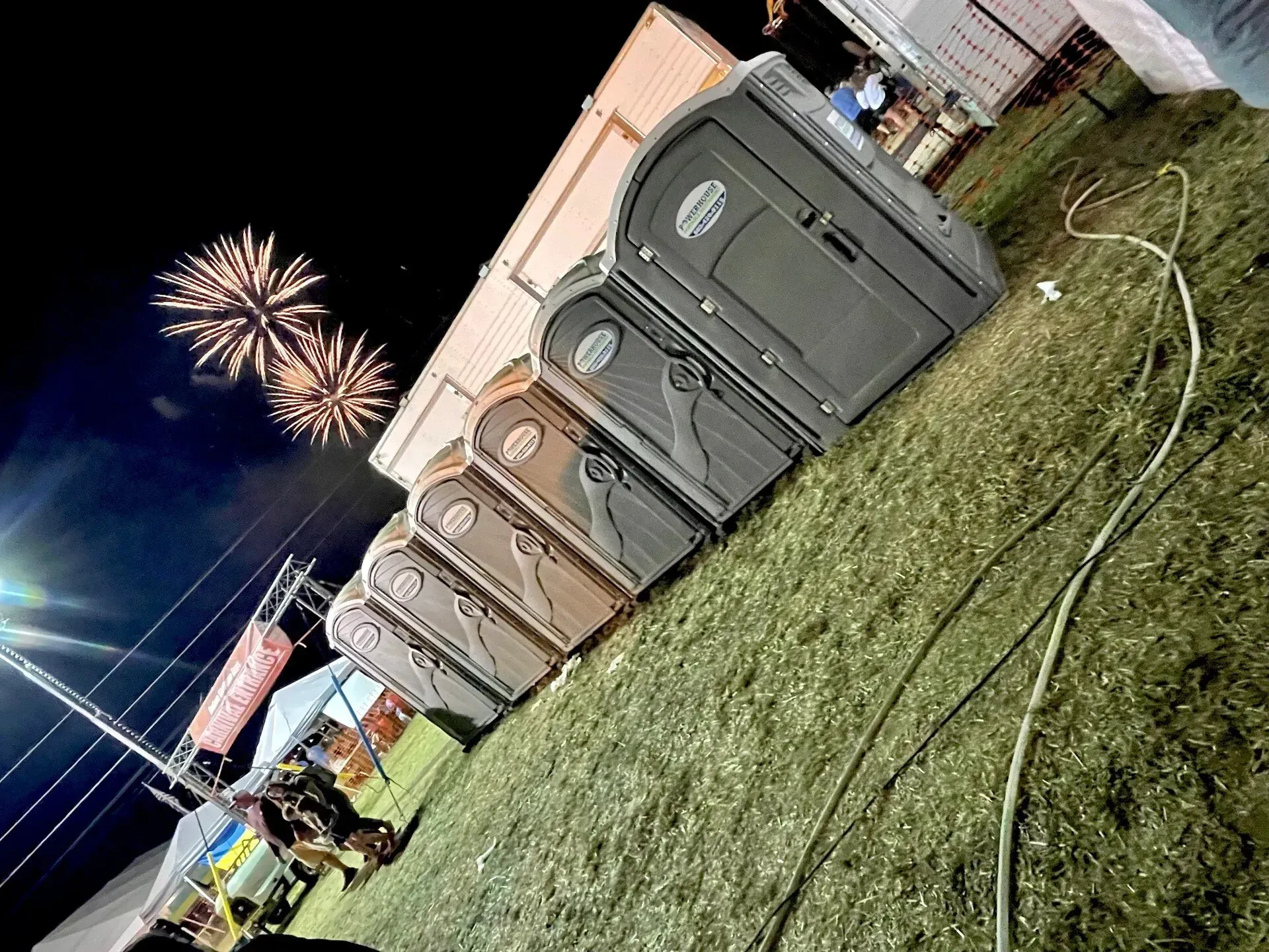 A row of portable toilets stands on a grassy field at night under fireworks.