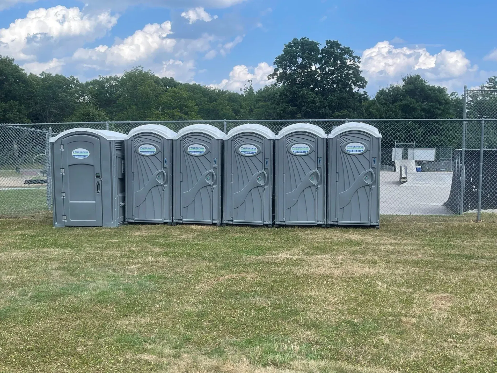 A row of six grey portable toilets lined up along a chain-link fence in a grassy outdoor area under a blue sky.