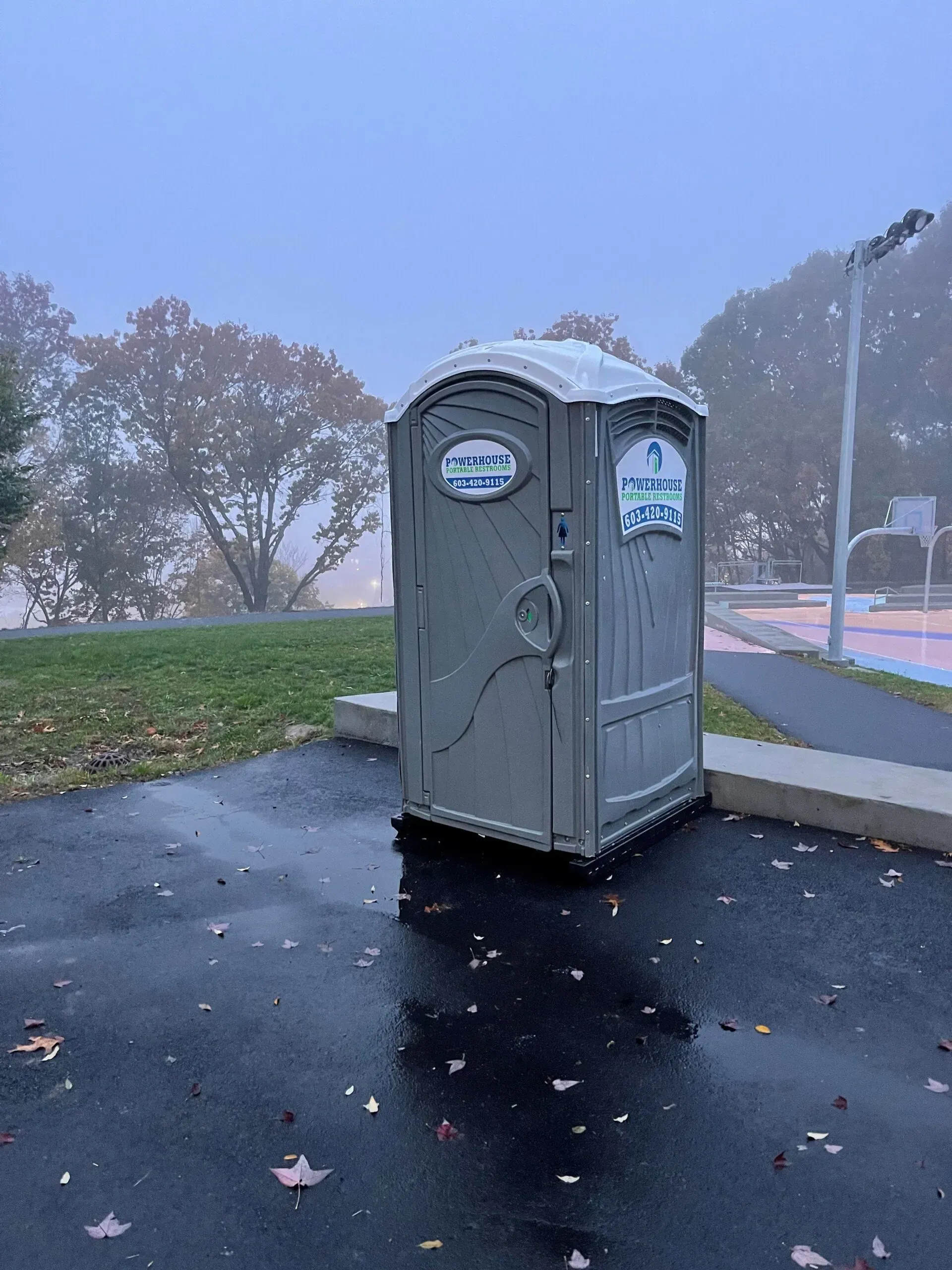 A tan portable toilet stands on a wet, paved lot in a park during a foggy, overcast morning.