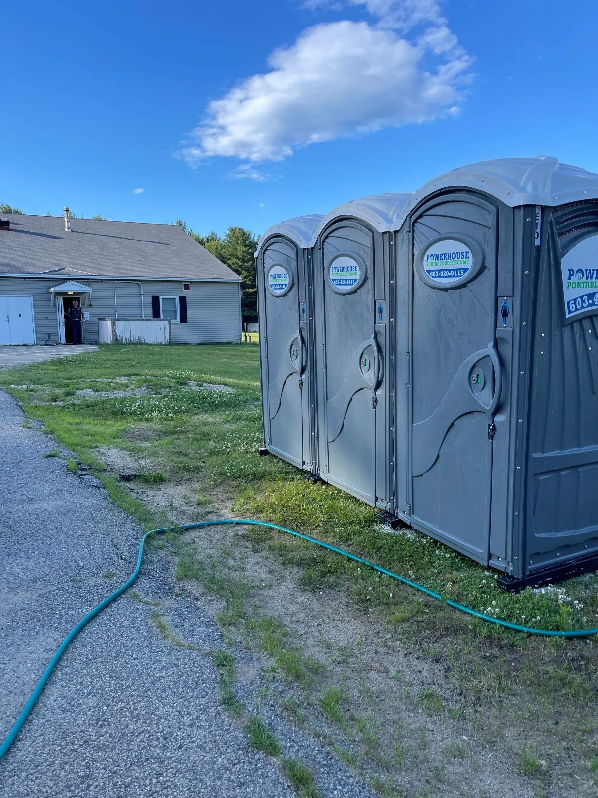 Three grey portable toilets sit on a grassy area next to a gravel driveway and a one-story house under a blue sky.