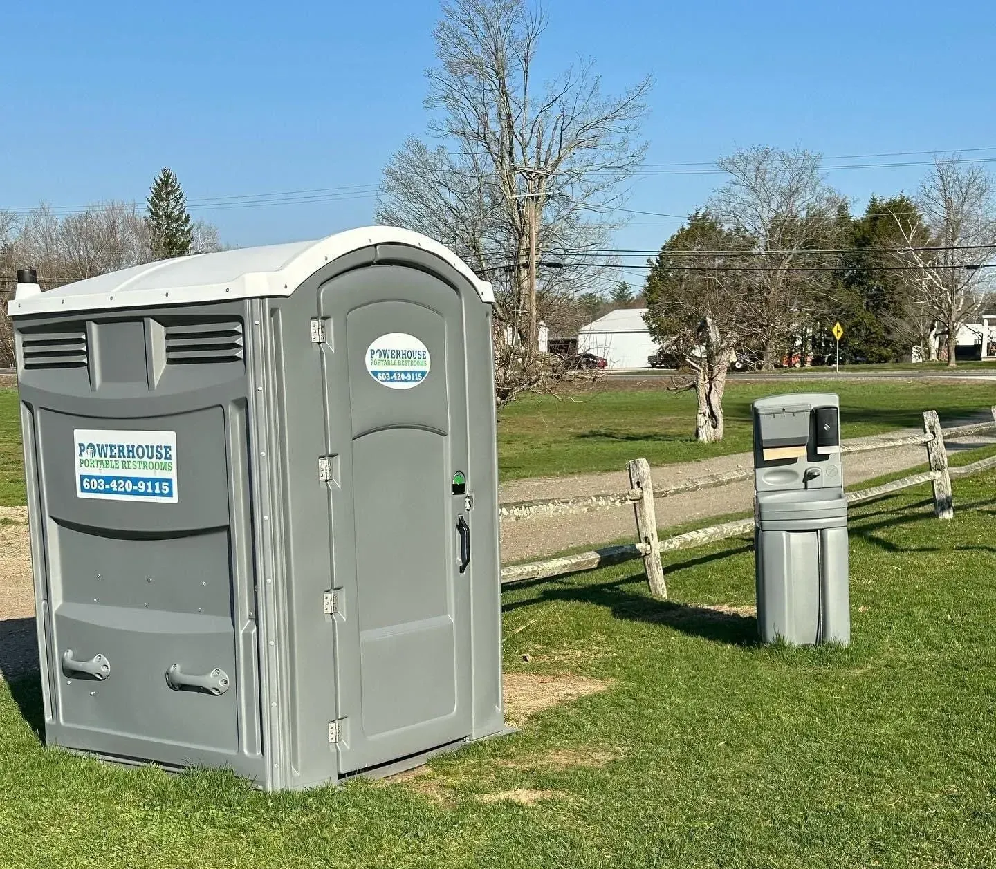 A gray portable toilet with a matching handwashing station set on a grassy field with a wooden fence.