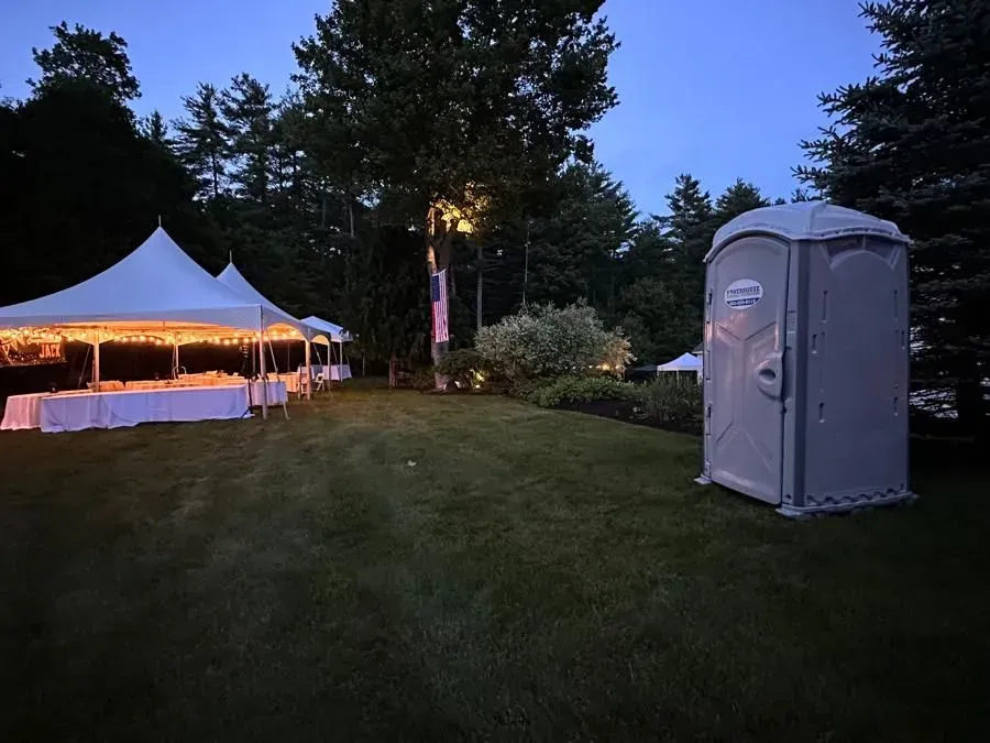 An outdoor event area at twilight, featuring white tents with interior lighting and a portable restroom on a lawn.