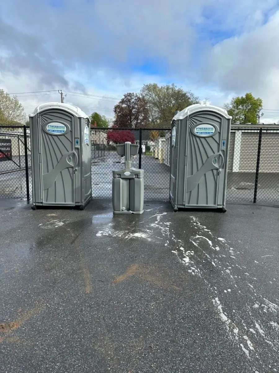 Two grey portable toilets and a handwashing station on an asphalt lot in front of a chain-link fence.