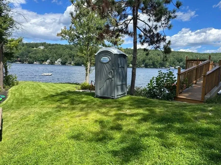 A gray portable toilet sits on a grassy lakeshore next to a wooden pier under a sunny sky with clouds.