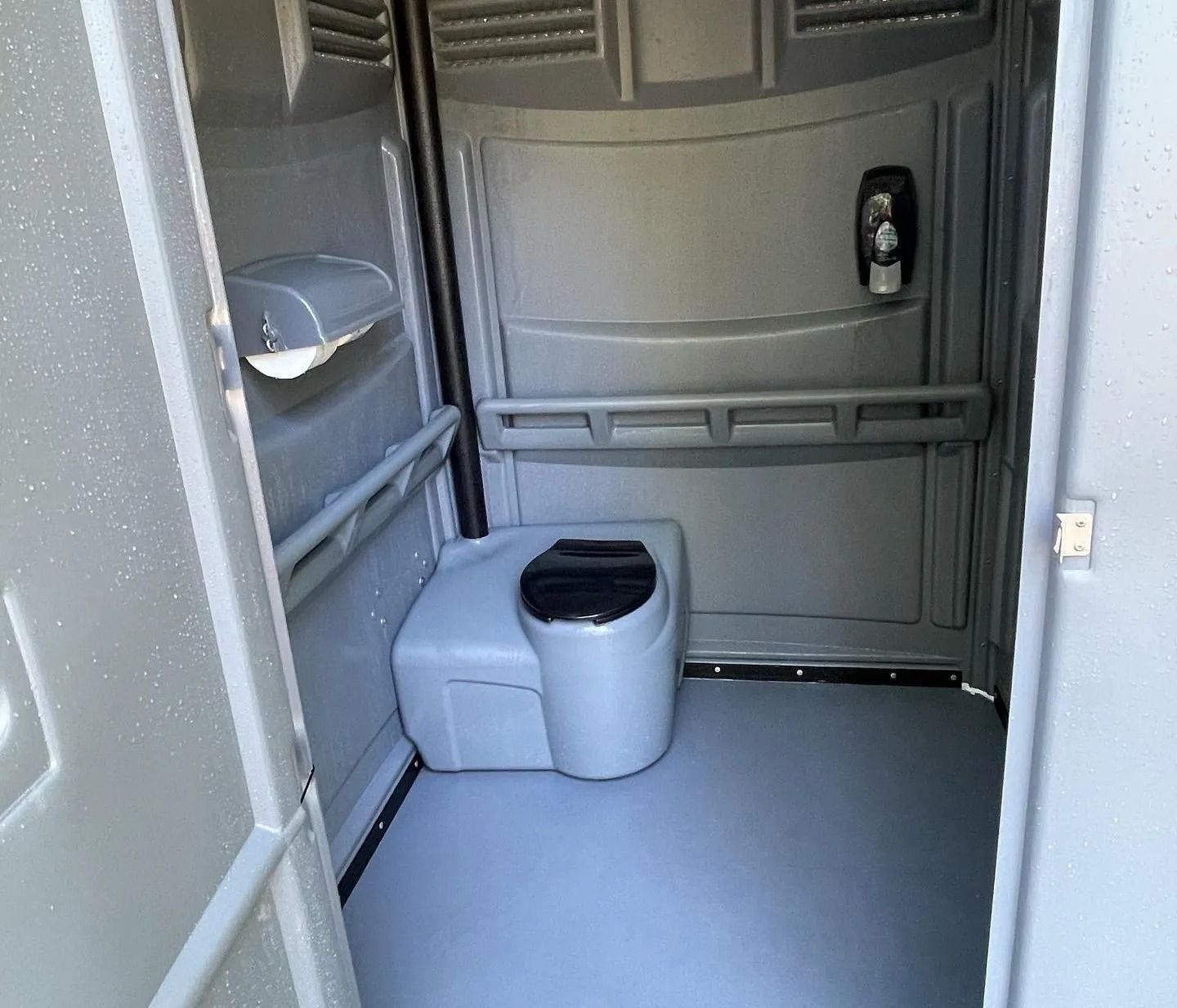The interior of a gray, plastic portable restroom featuring a black toilet seat, handrails, and a soap dispenser.
