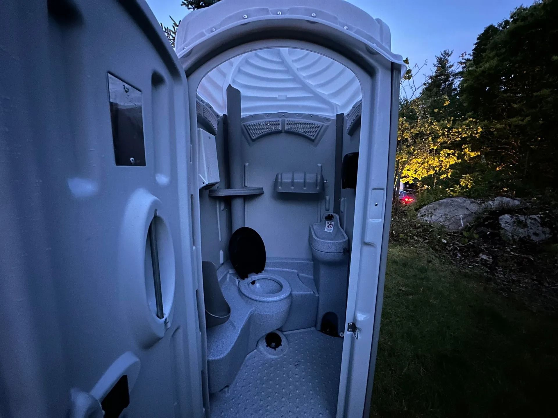 An open door reveals the interior of a grey portable toilet, featuring a sink, a toilet, and shelves in a grassy setting.