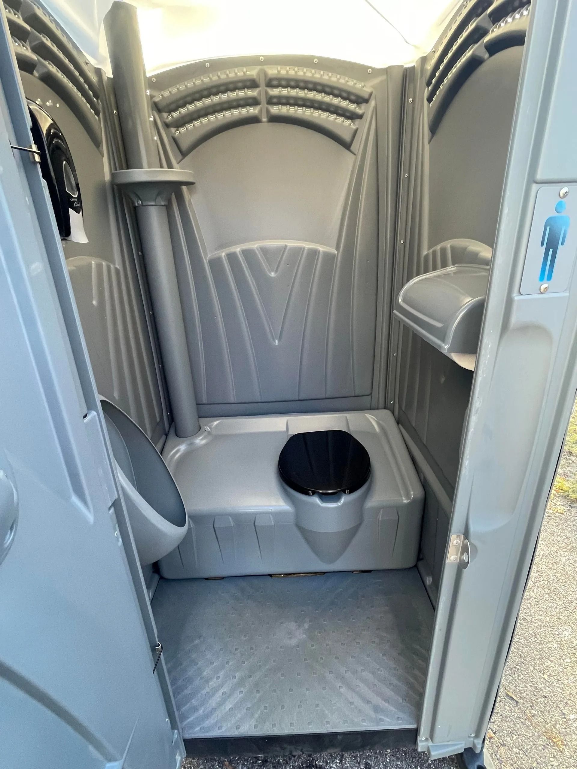 An interior view of a gray portable toilet featuring a black toilet seat, a wall-mounted urinal, and a toilet paper holder.