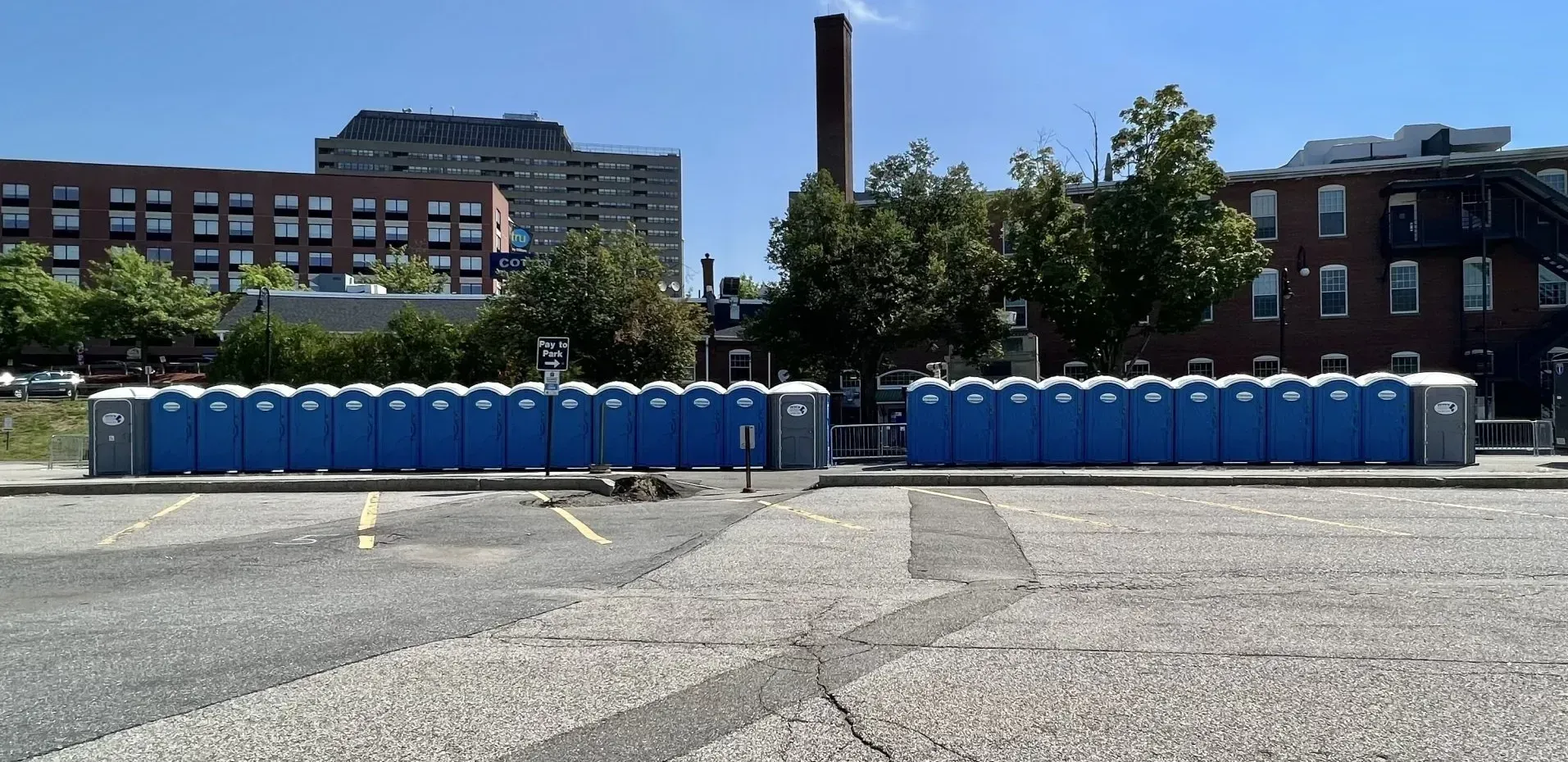 Two rows of blue portable toilets sit in a gravel parking lot against a backdrop of brick buildings and a blue sky.