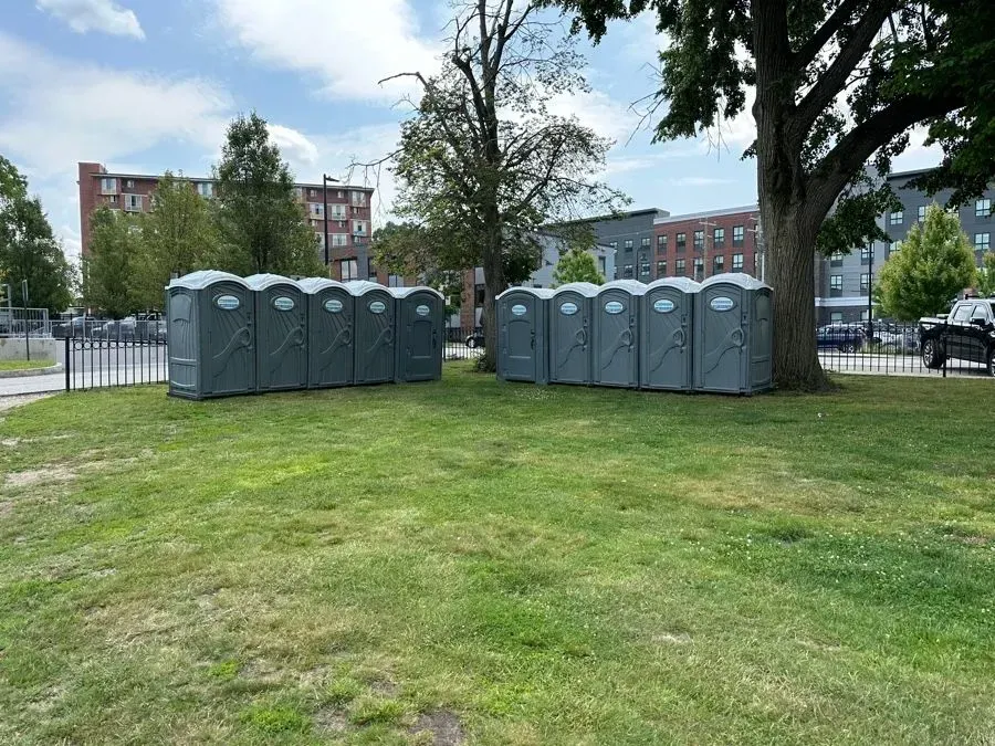 A row of grey portable toilets sits on a grassy field near a tree and buildings under a bright, cloudy sky.