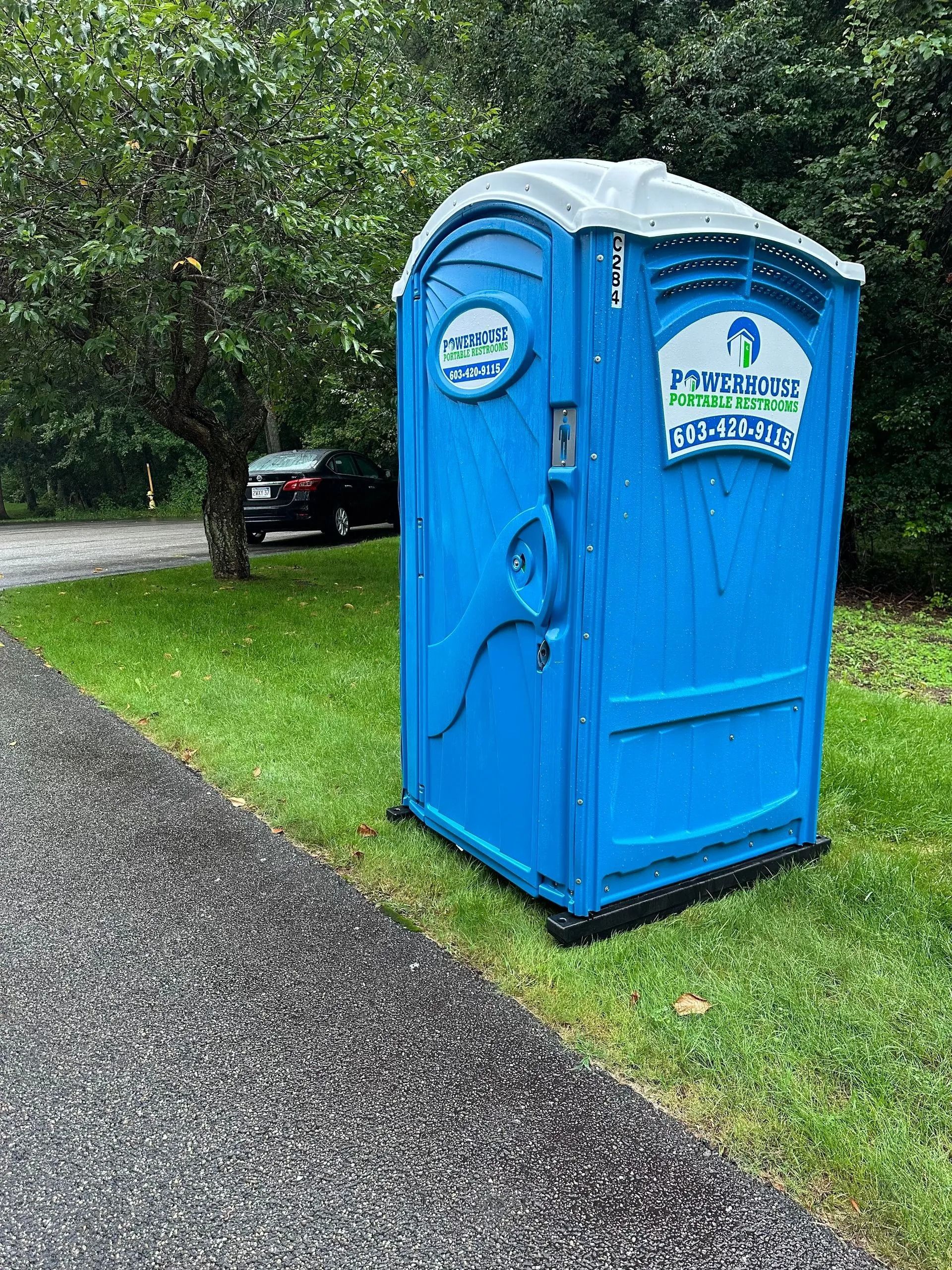 A blue portable restroom unit stands on a grassy area next to a gravel path, with trees and a parked car in the background.