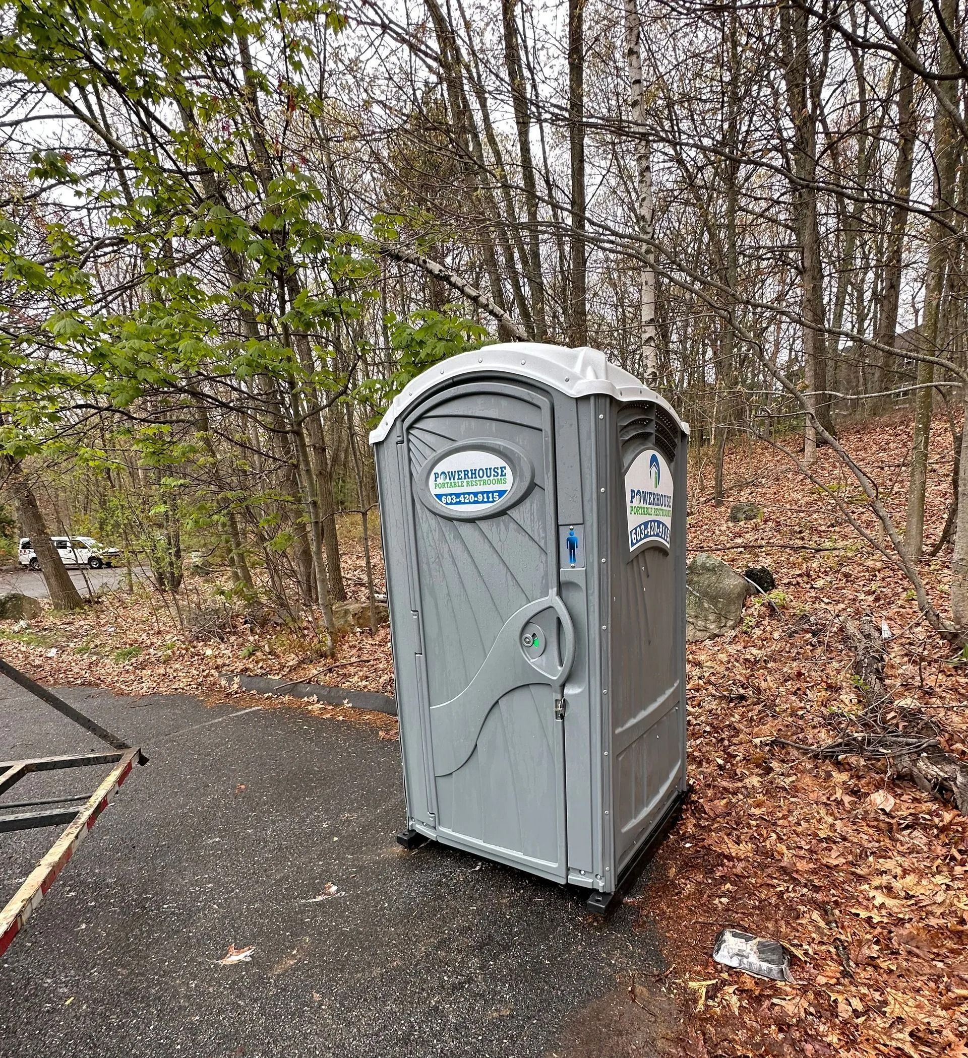 A grey portable toilet stands on a paved surface at the edge of a wooded area covered in fallen leaves.