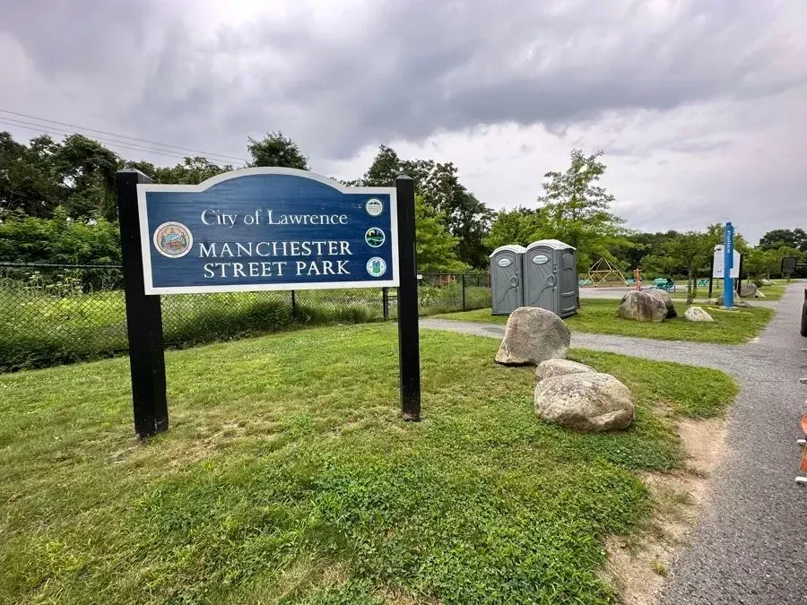 A blue City of Lawrence Manchester Street Park sign stands on a grassy lot with portable toilets and gravel paths.